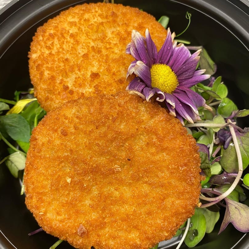 Two golden-brown fried patties on a bed of fresh greens, garnished with a purple flower.